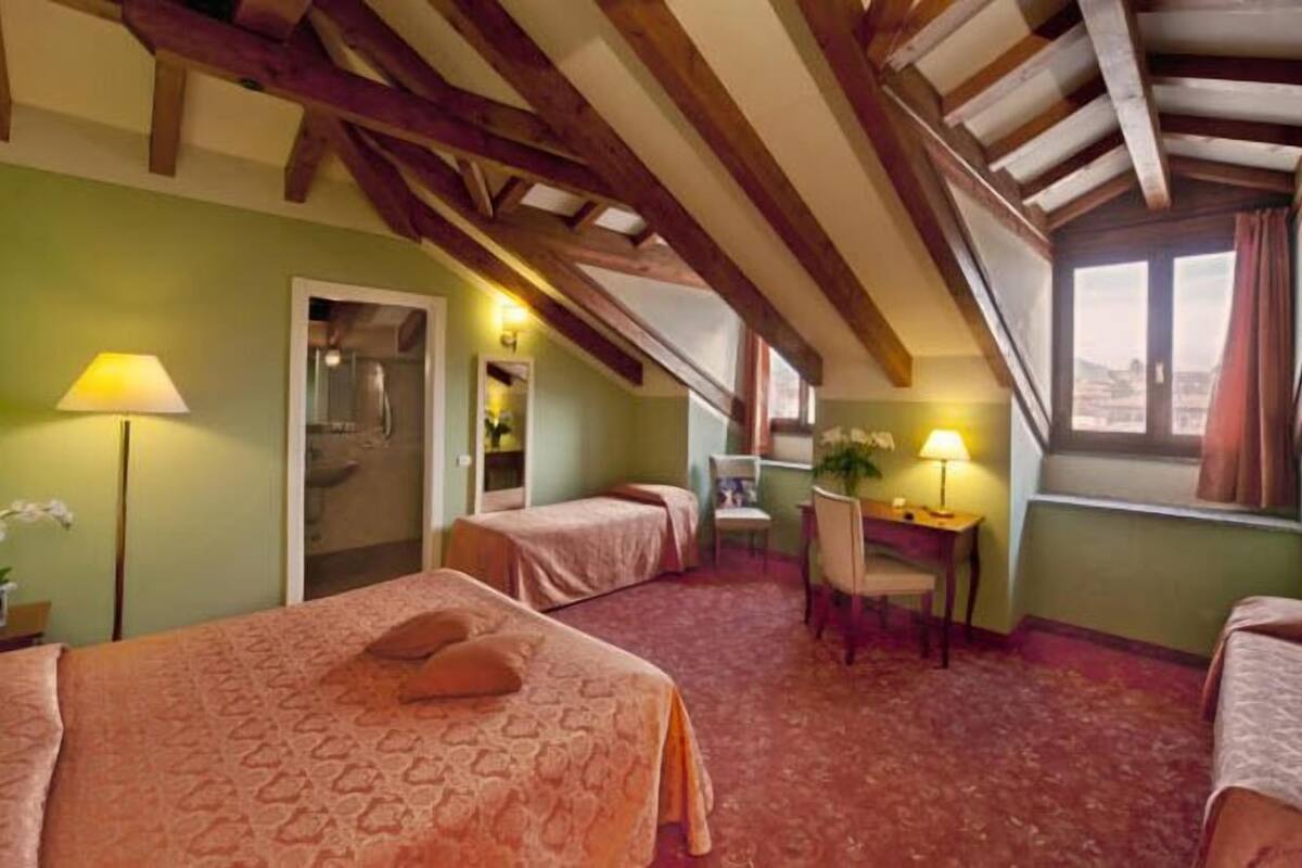 A room with two beds, a desk, and a chair. There is a window with curtains, a lamp, and a plant.