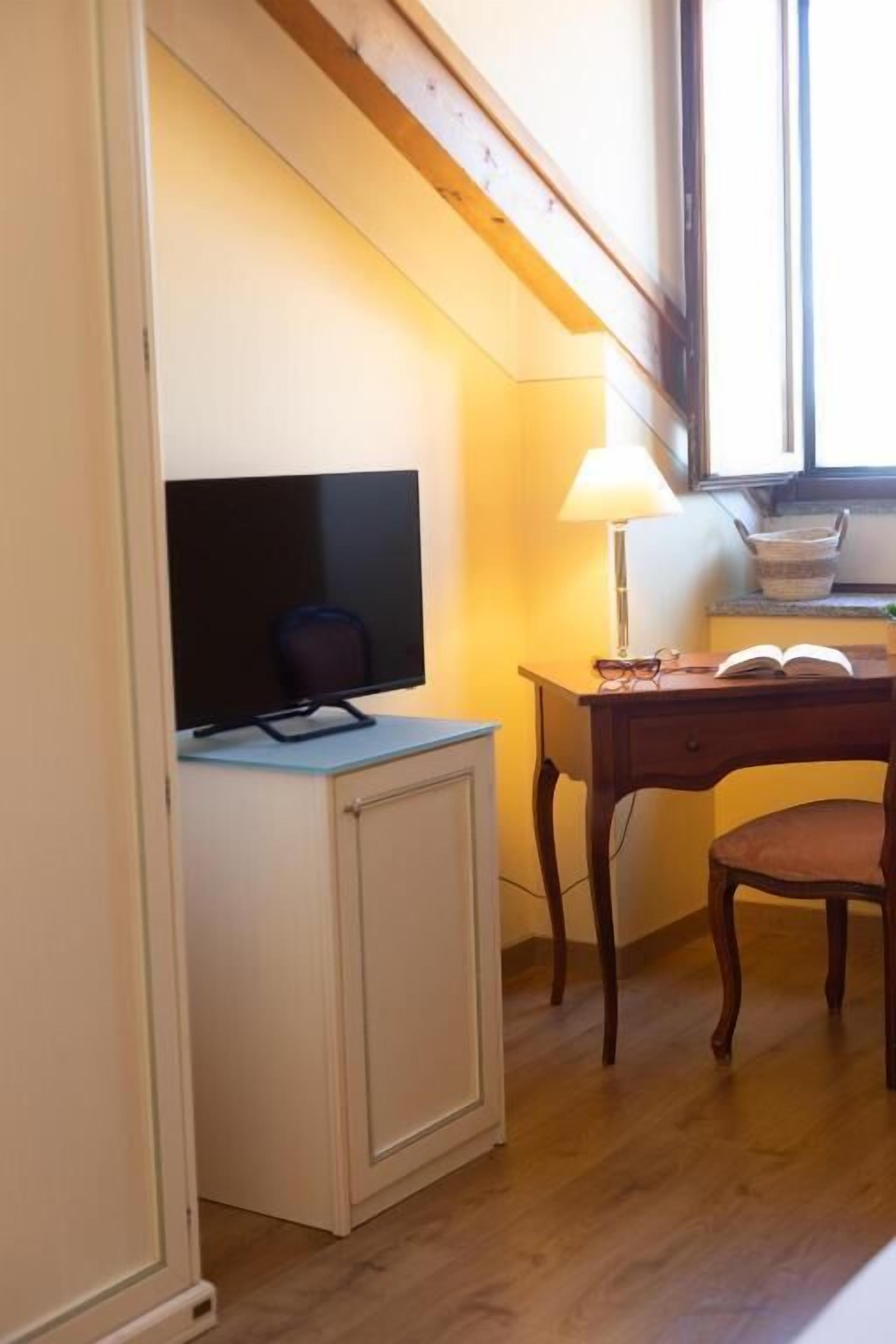 Een kamer met een houten bureau, een stoel, een televisie op een kast, een lamp en een raam met natuurlijk licht.