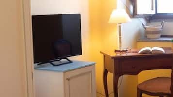 Een kamer met een houten bureau, een stoel, een televisie op een kast, een lamp en een raam met natuurlijk licht.