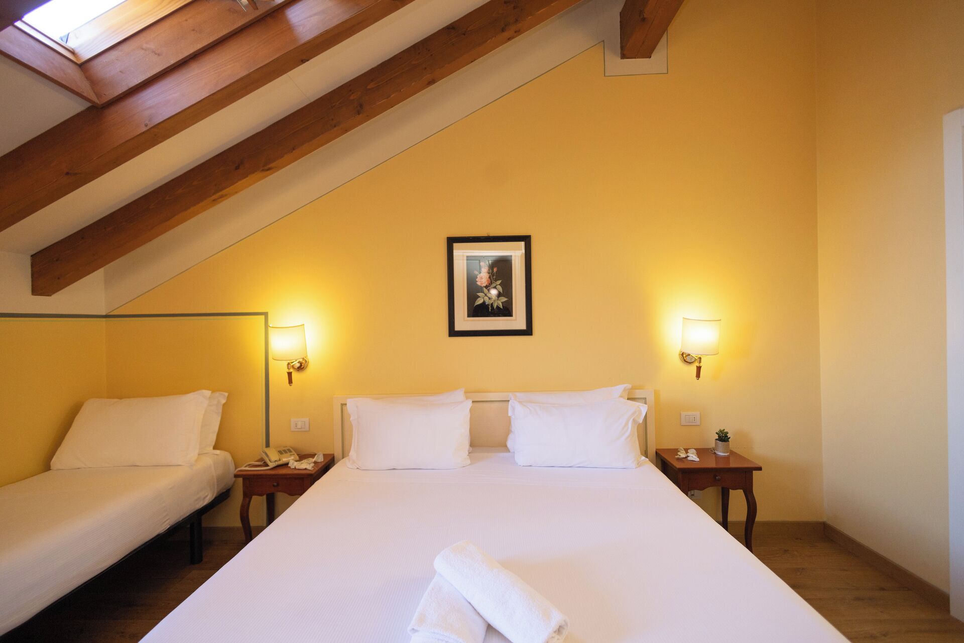Ein Hotelzimmer mit hoher Decke, Holzbalken, zwei Einzelbetten, einem großen Bett, Nachttischlampen, einem gerahmten Bild an der Wand und einem kleinen Tisch mit einer Pflanze.