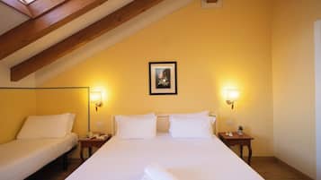 Ein Hotelzimmer mit hoher Decke, Holzbalken, zwei Einzelbetten, einem großen Bett, Nachttischlampen, einem gerahmten Bild an der Wand und einem kleinen Tisch mit einer Pflanze.
