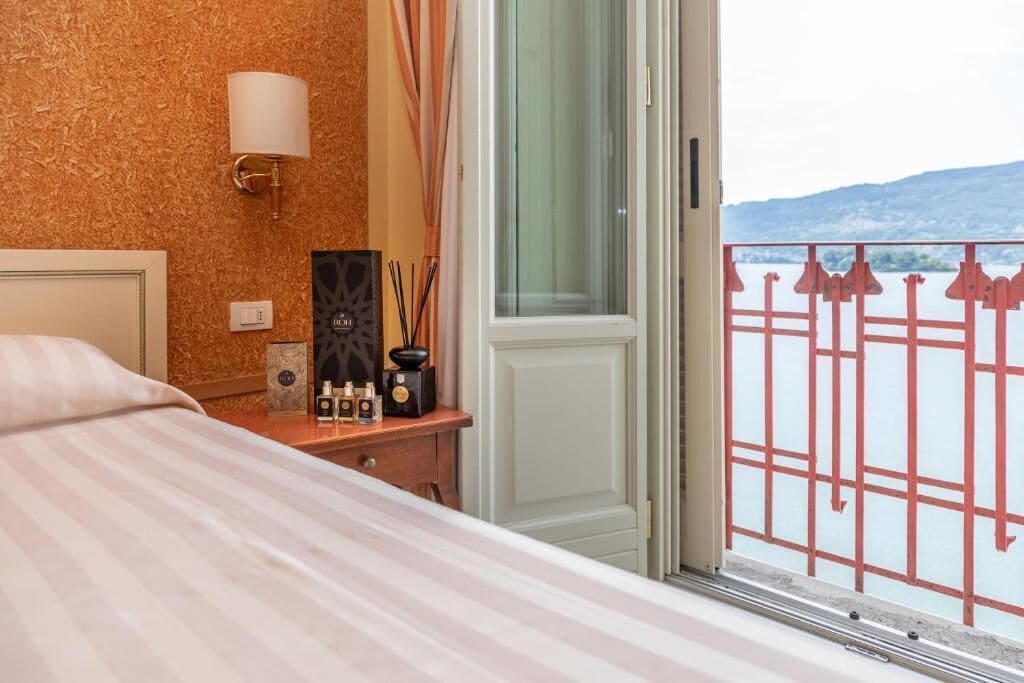 Habitación de hotel con cama, mesita de noche con lámpara, ventana con vista a un cuerpo de agua y barandilla.