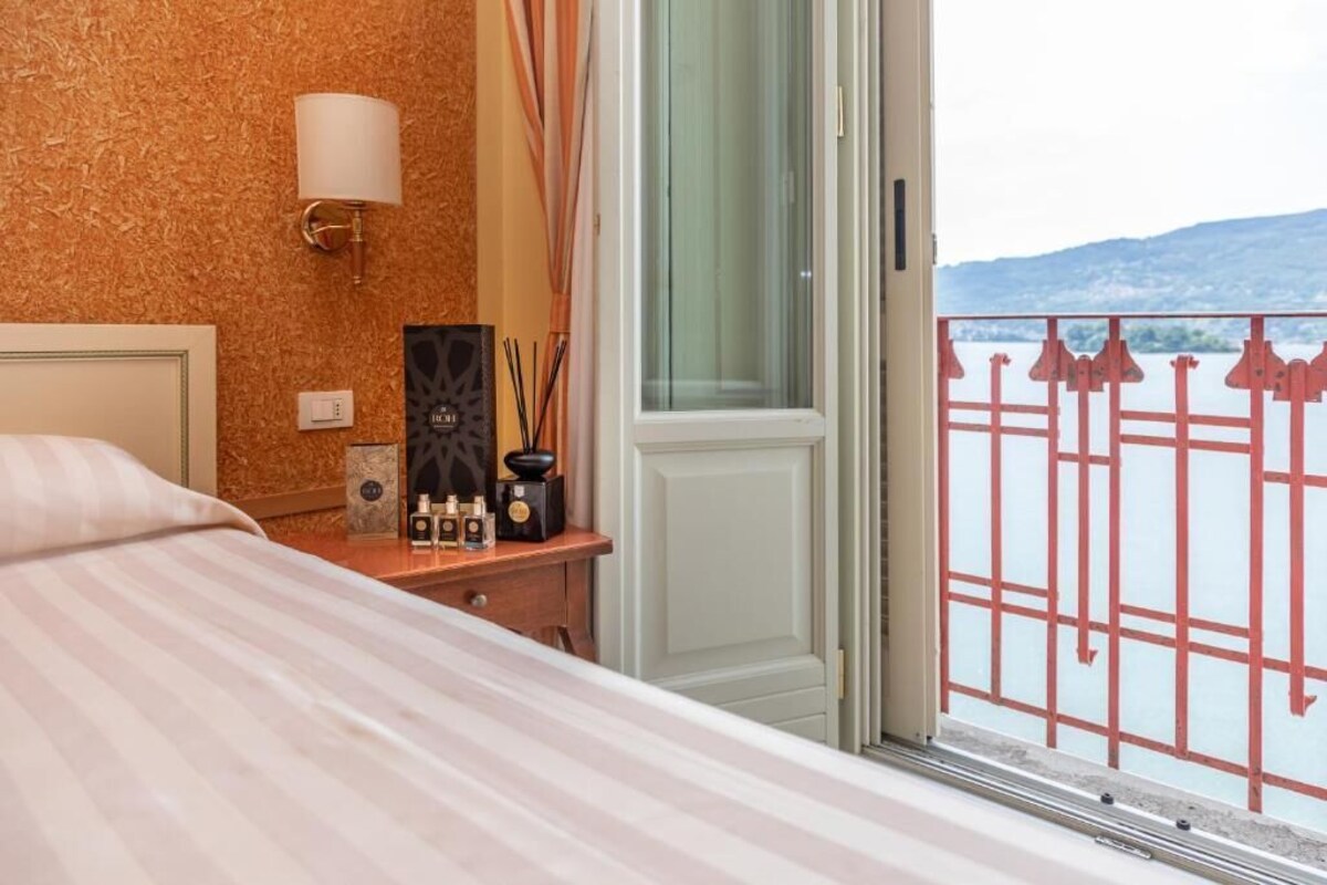 A hotel room with a bed, a nightstand with a lamp, a window with a view of a body of water and a railing.