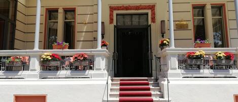 Entrada de hotel con alfombra roja que lleva a una puerta negra, columnas blancas y macetas con flores en los balcones.