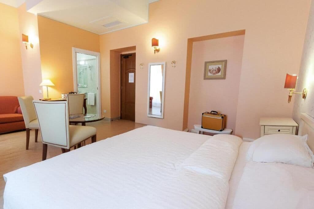 Une chambre d’hôtel avec un grand lit, un bureau avec une chaise, une petite table, une valise et une table de chevet.