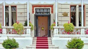 Entrada de hotel con alfombra roja que conduce a una puerta, flanqueada por columnas blancas y plantas en macetas.