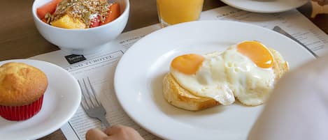 Un desayuno con un muffin, un plato con huevo, un bol de fruta, un vaso de zumo de naranja y una taza de café sobre una mesa.