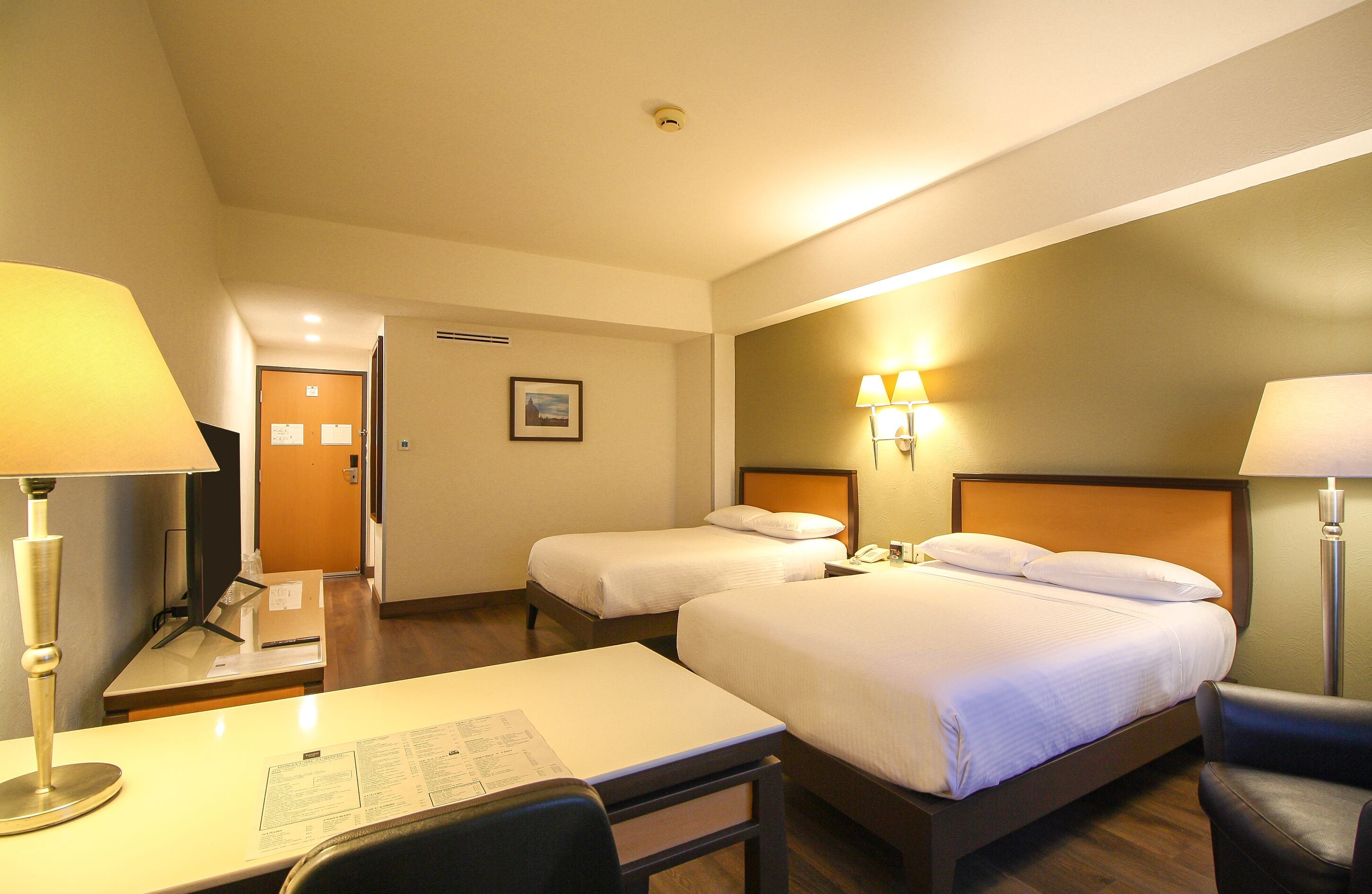 A hotel room with two beds, a desk with a lamp, a chair, and a bedside table.