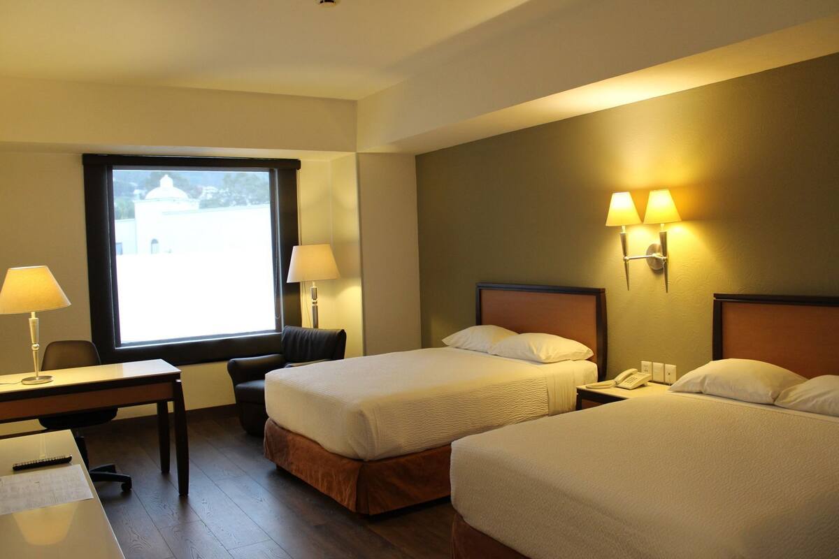 A hotel room with two beds, a desk, a chair, and a window with a view.