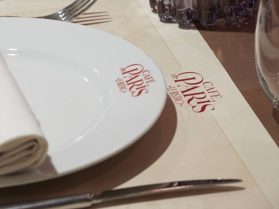 Een netjes gedekte tafel met een wit bord, gevouwen servet en bestek. De tafelkleed heeft 'Café Paris' erop staan.