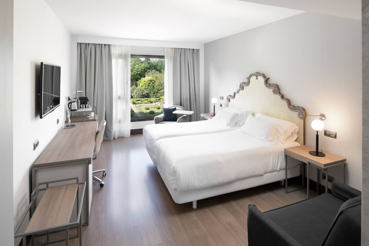 A hotel room with a large bed, a desk, a chair, a TV, and a view of a garden.