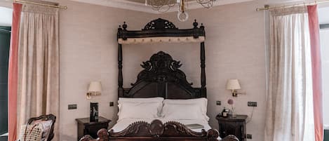 Un dormitorio con una cama grande y ornamentada, dos mesitas de noche con lámparas, una silla y un candelabro.