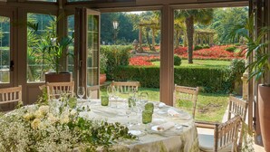 Un comedor con una mesa dispuesta para una comida, rodeada de sillas, y vista a un jardín con flores y follaje.