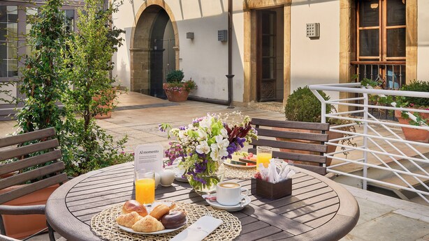 Un patio al aire libre con una mesa redonda preparada para el desayuno, que incluye cruasanes, una bebida y un menú.