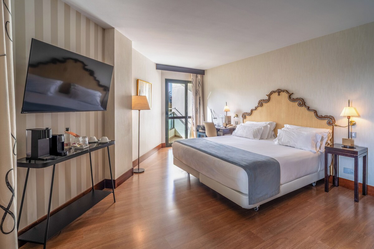 A hotel room with a large bed, a flat-screen TV mounted on the wall, a desk with a coffee maker, and a view of the outside through a glass door.