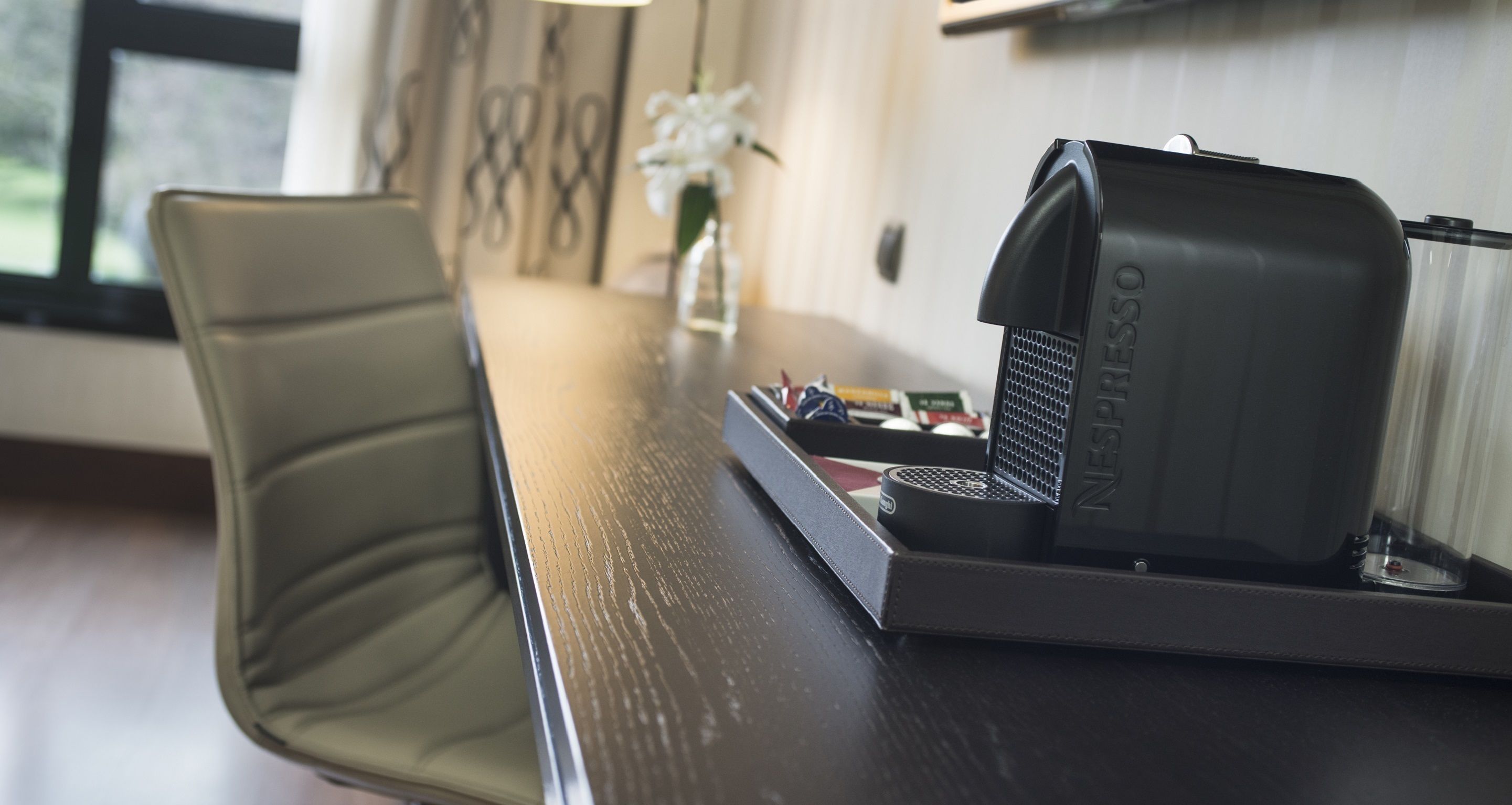 A Nespresso coffee machine on a wooden table in a room with a chair and a vase of flowers.