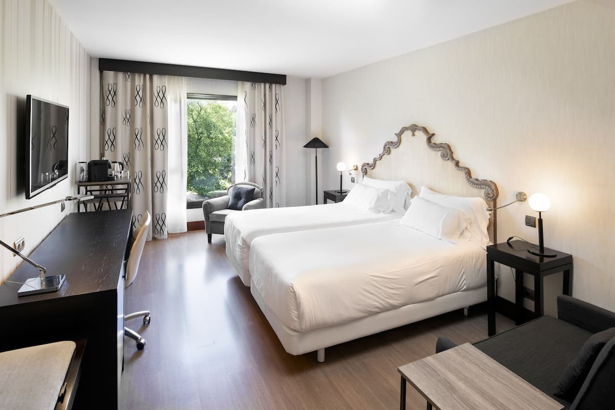 A hotel room with two beds, a desk, a chair, a TV, and a window with curtains.