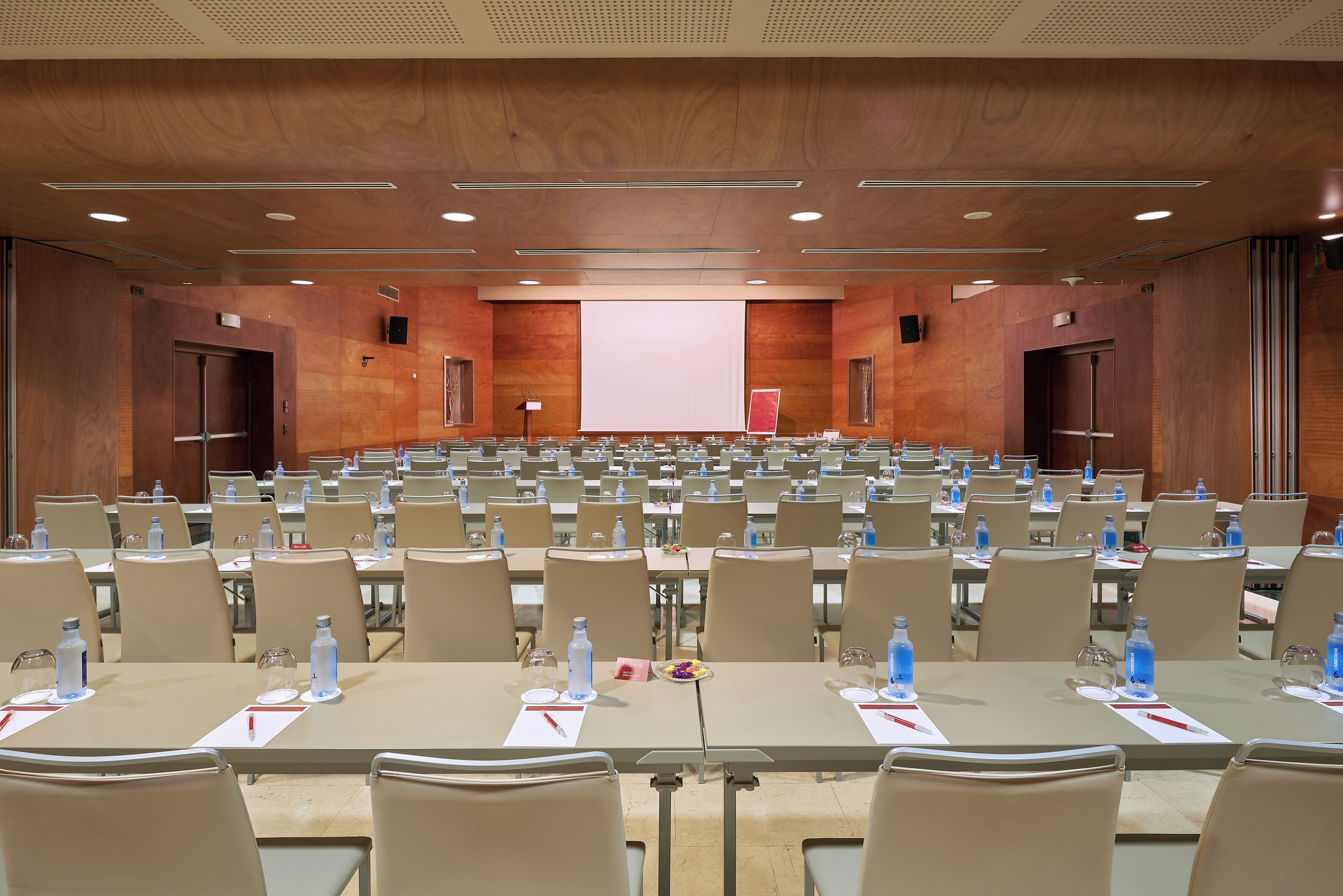 Sala de conferências com mesas e cadeiras dispostas em fileiras, equipada com garrafas de água e placas de identificação.