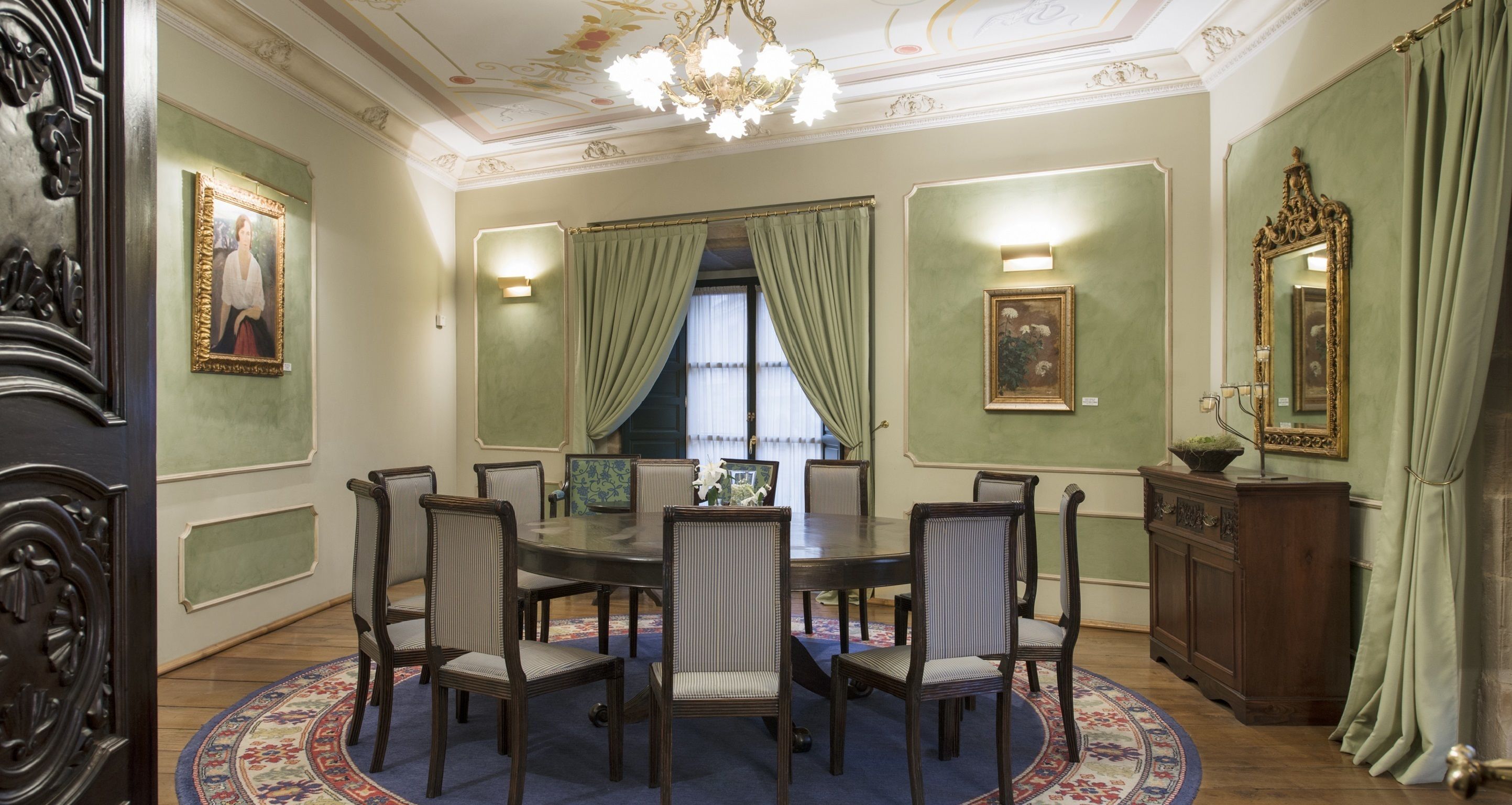 Ein formeller Speisesaal mit einem runden Tisch, sechs Stühlen, einer Chandelüster und grünen Vorhängen.