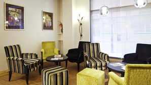 Een kamer met gestreepte fauteuils, een gele fauteuil en een zwarte bank. Twee ingelijste kunstwerken hangen aan de muur, er staat een kleine tafel met een vaas en een raam met jaloezieën.