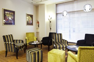 A room with striped armchairs, a yellow armchair, and a black sofa. There are two framed artworks on the wall, a small table with a vase, and a window with blinds.