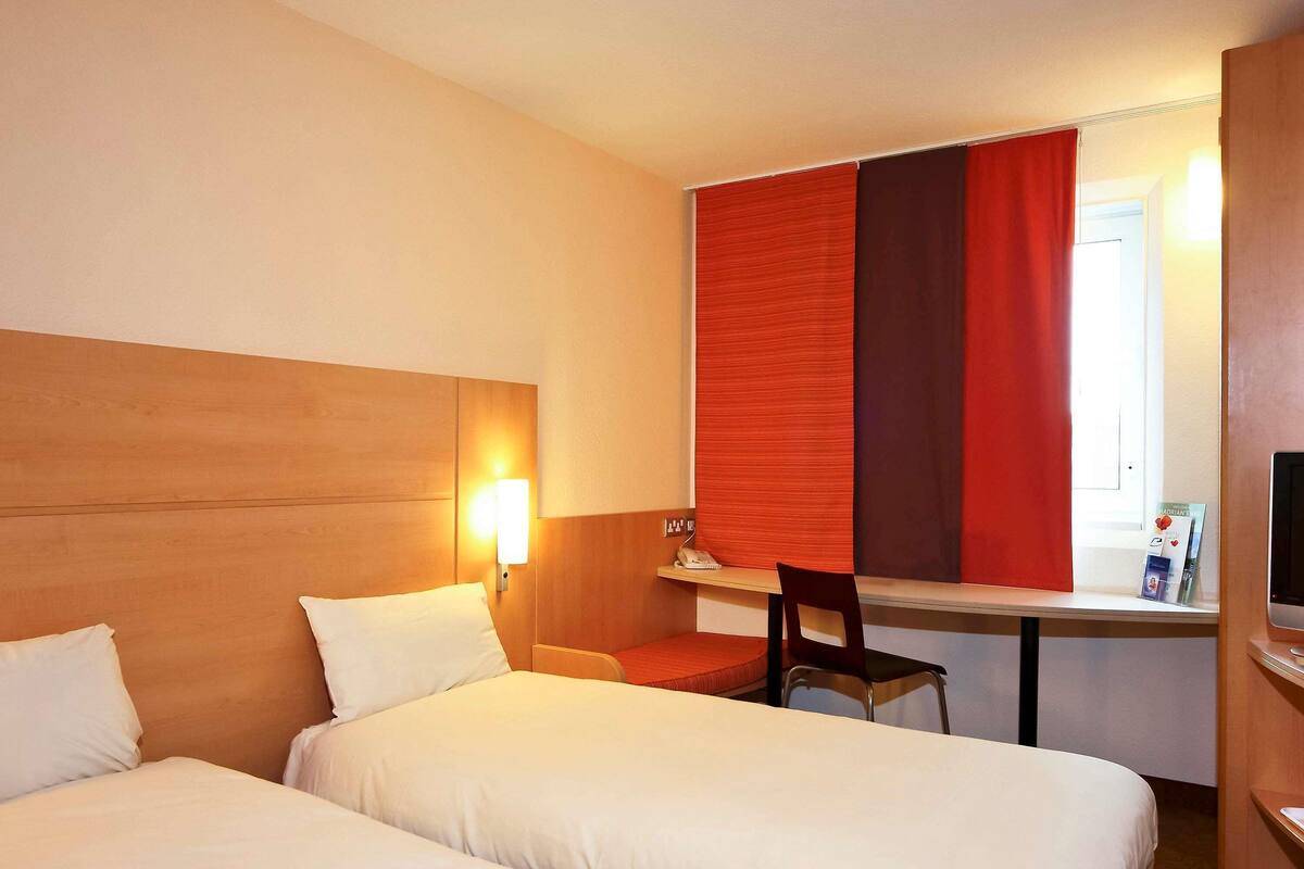 Habitación de hotel con dos camas, un escritorio, una silla, un televisor y una ventana con cortinas rojas.