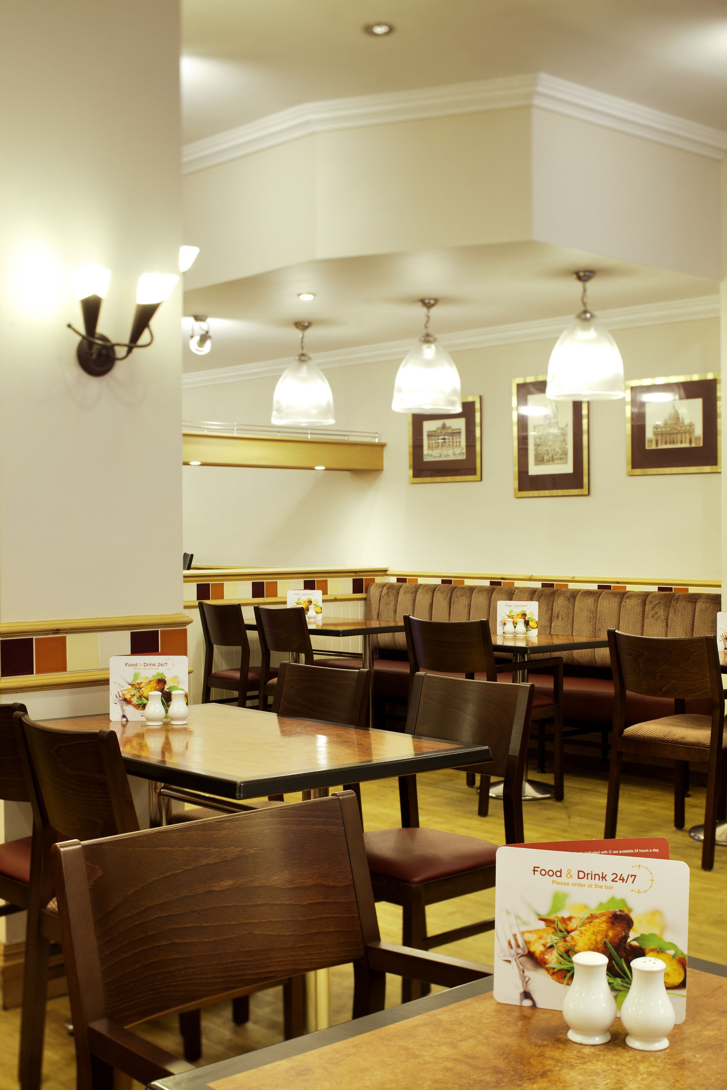Restaurante com mesas e cadeiras de madeira, quadros nas paredes e um aviso indicando serviço de alimentação e bebidas 24 horas por dia.