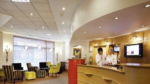 Een hotel receptie met een gebogen houten balie, een televisiescherm en een zithoek met gestreepte stoelen en een gele bank.