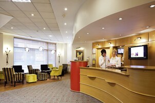 A hotel reception area with a curved wooden reception desk, a television screen, and a seating area with striped chairs and a yellow sofa.