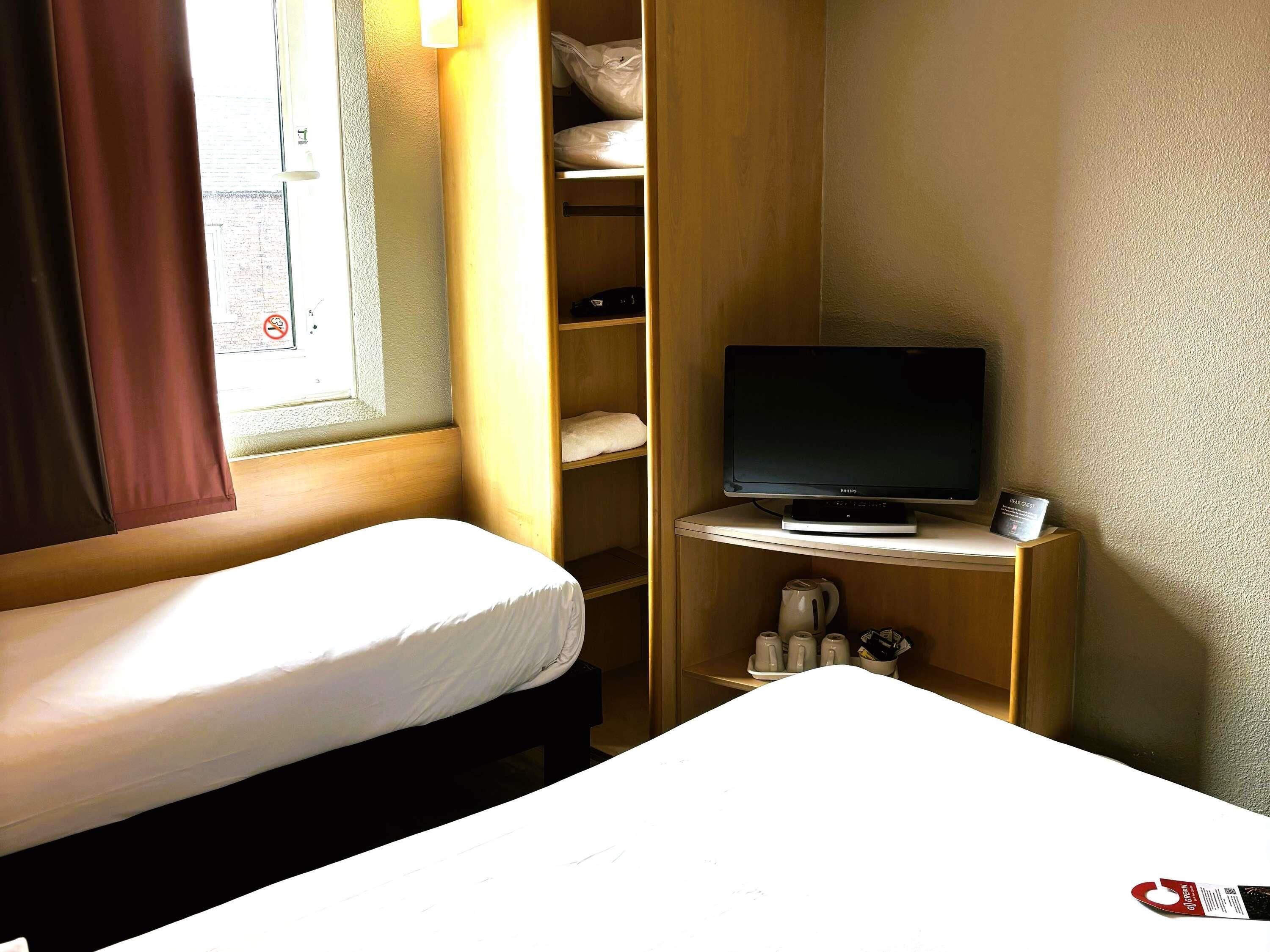 A small hotel room with a single bed, a television on a stand, a shelf with toiletries, and a window with curtains.