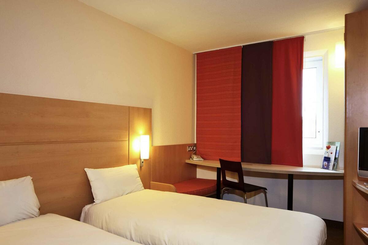 Ein Hotelzimmer mit zwei Betten, einem Schreibtisch, einem Stuhl, einem Fernseher und einem Fenster mit roten Vorhängen.