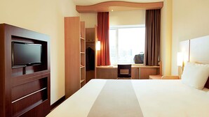 Een hotelkamer met een groot bed, een televisie bevestigd aan een houten kast, een bureau met een stoel, en een raam met gordijnen.