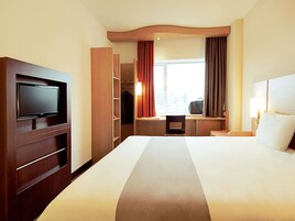 A hotel room with a large bed, a television mounted on a wooden cabinet, a desk with a chair, and a window with curtains.