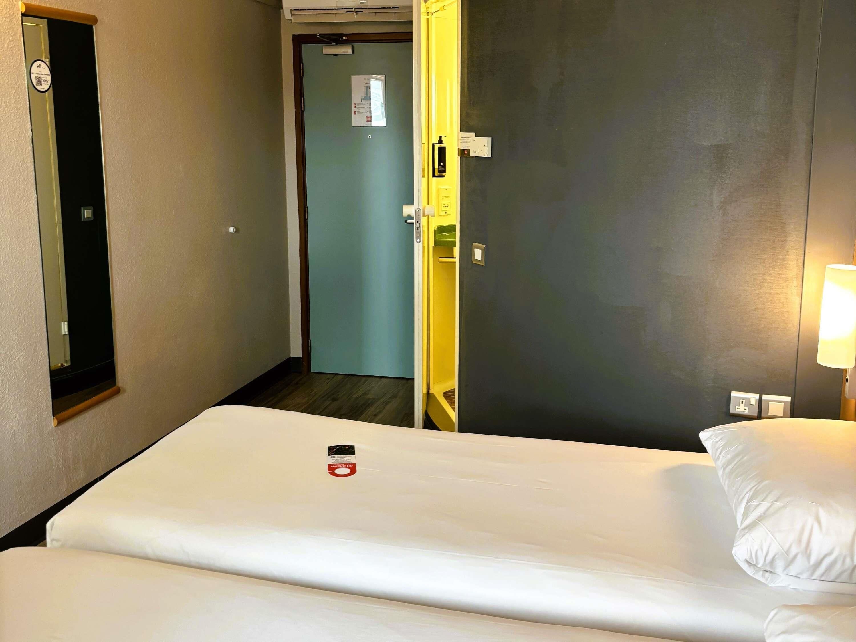 A hotel room with a bed, a nightstand with a lamp, a door, and a mirror.