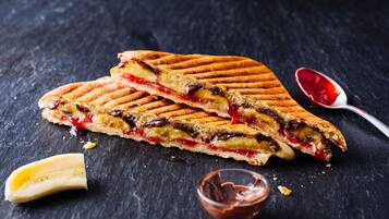 Un sándwich a la plancha con relleno visible, una cuchara con mermelada y un tazón pequeño de crema de chocolate sobre una superficie oscura.