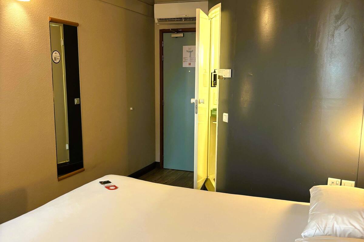 A hotel room with a bed, a mirror, a door, and a wall-mounted air conditioning unit.