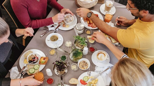 Een groep mensen dineert aan een tafel met diverse gerechten, waaronder roerei, soep en brood.