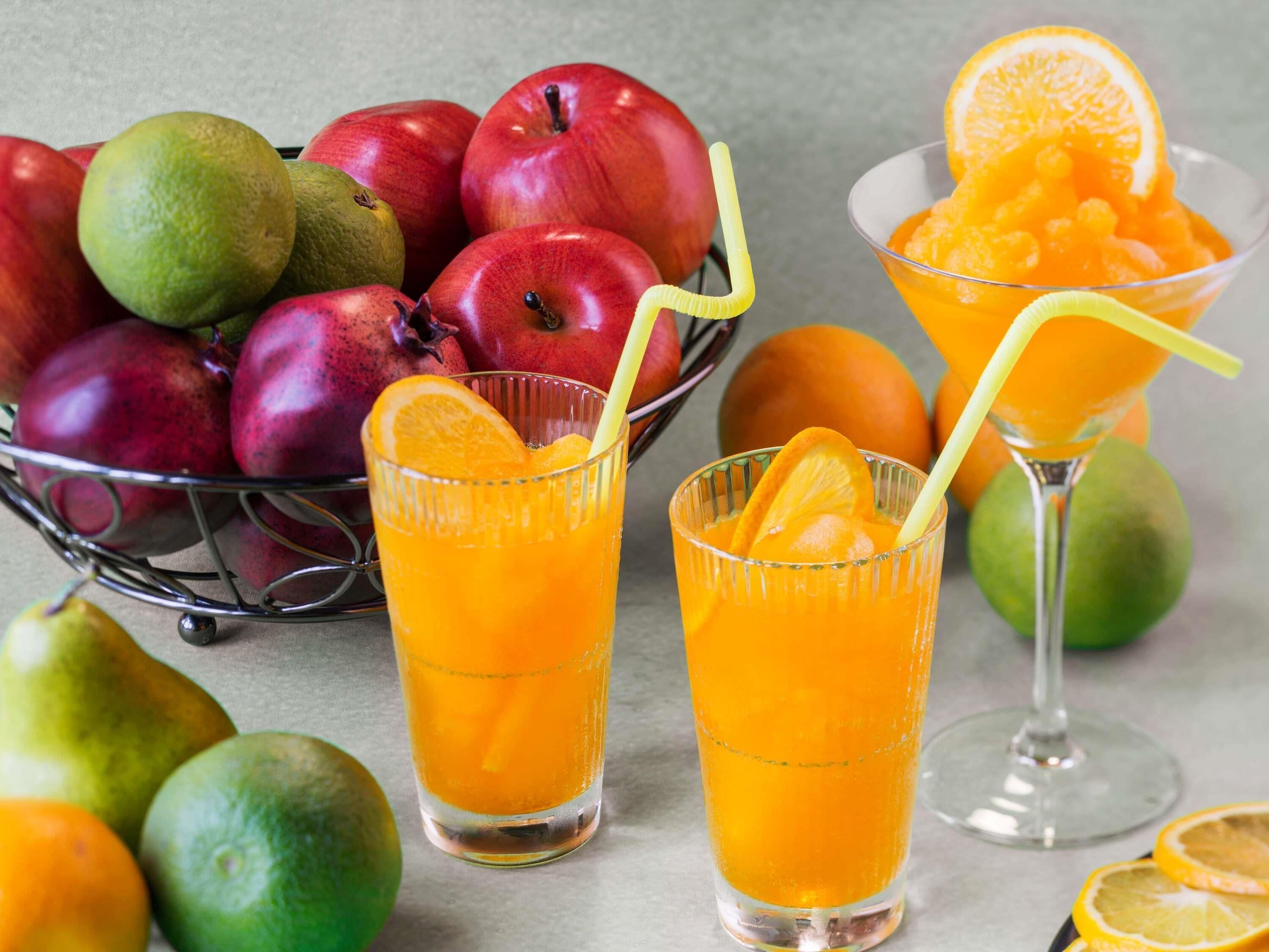 Een schaal met appels en sinaasappels, en twee glazen sinaasappelsap met rietjes.