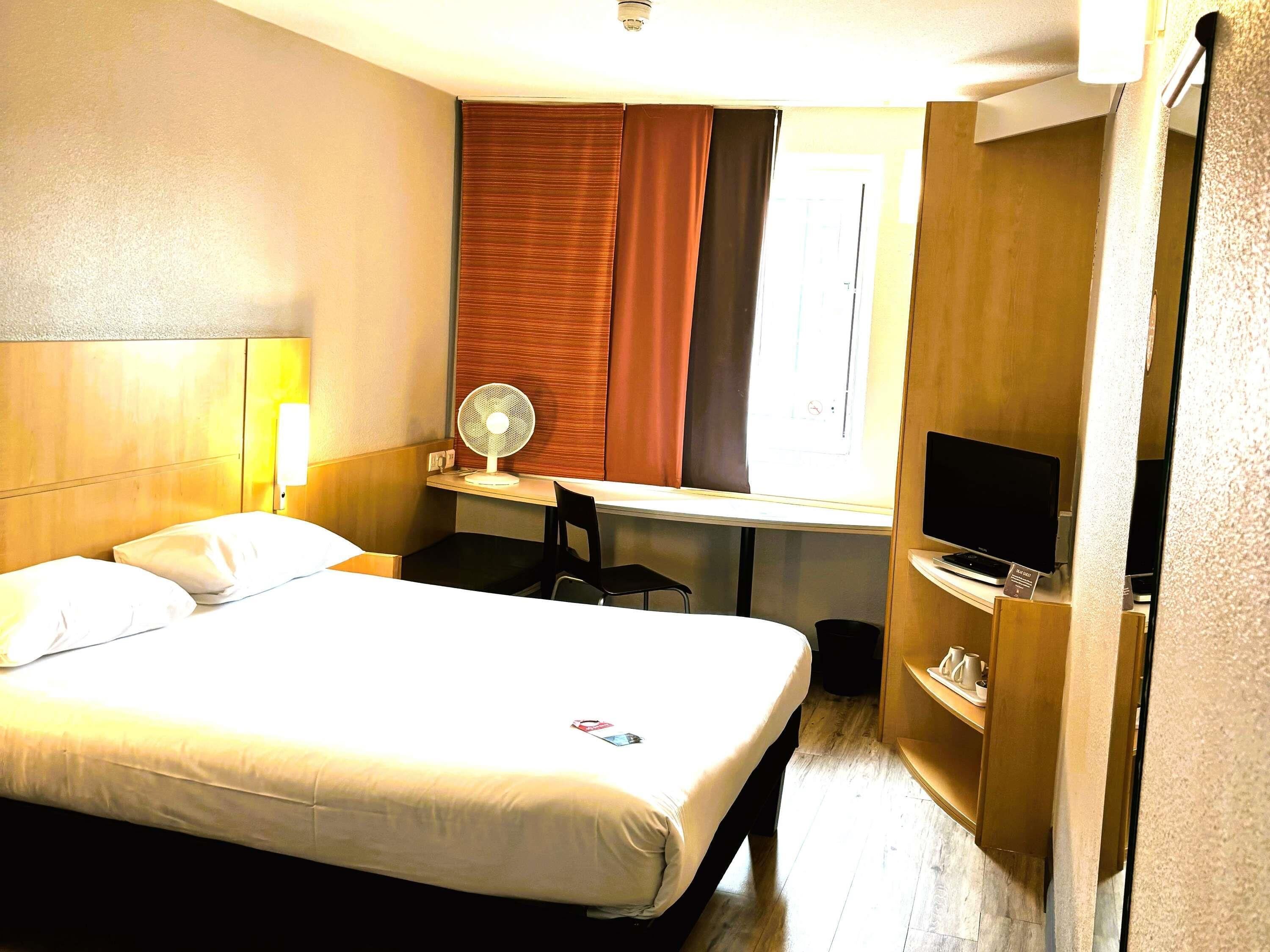 Habitación de hotel con cama, escritorio, silla, televisión y ventana con cortinas.