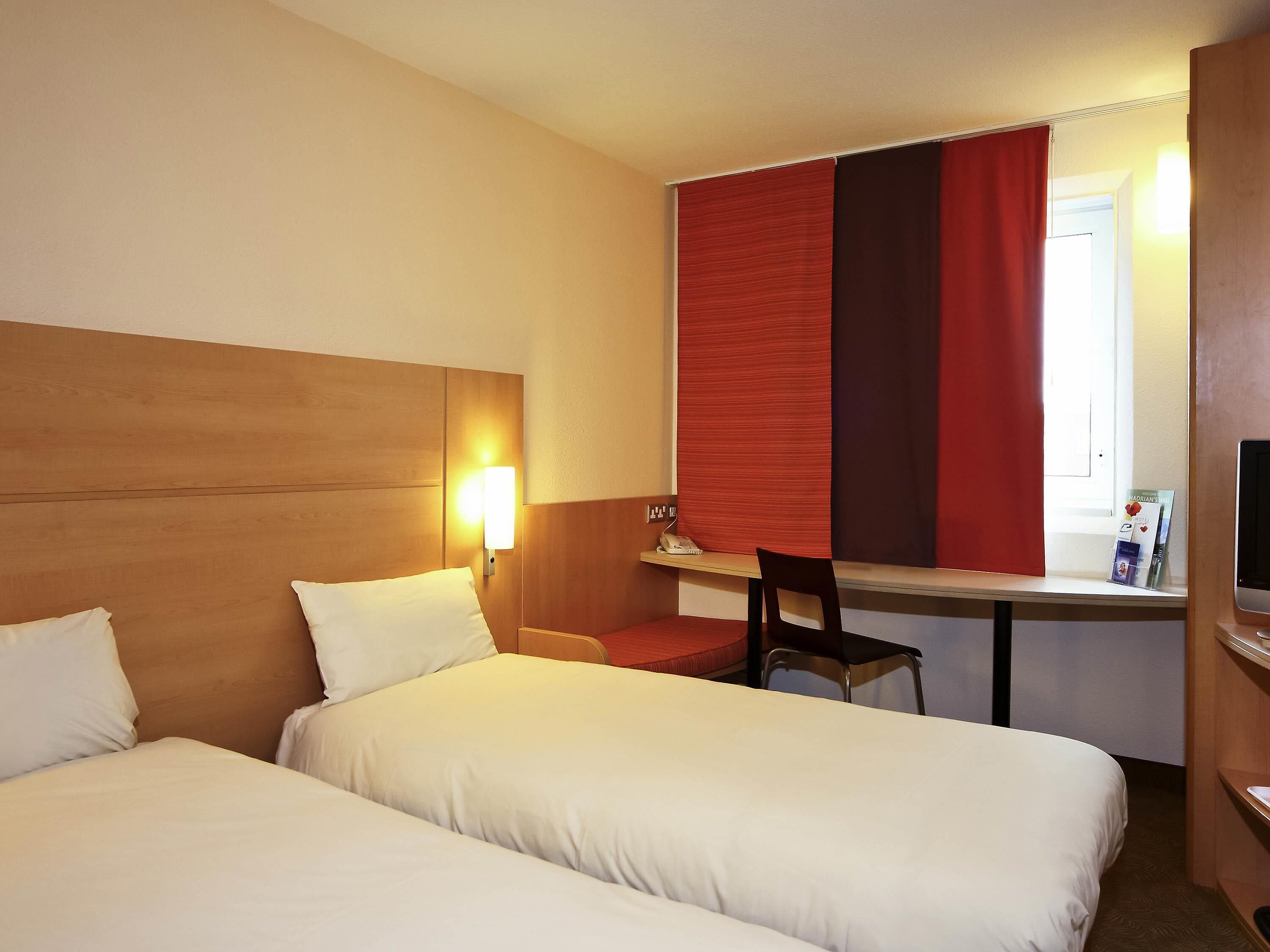 A hotel room with two beds, a desk, a chair, a TV, and a window with red curtains.