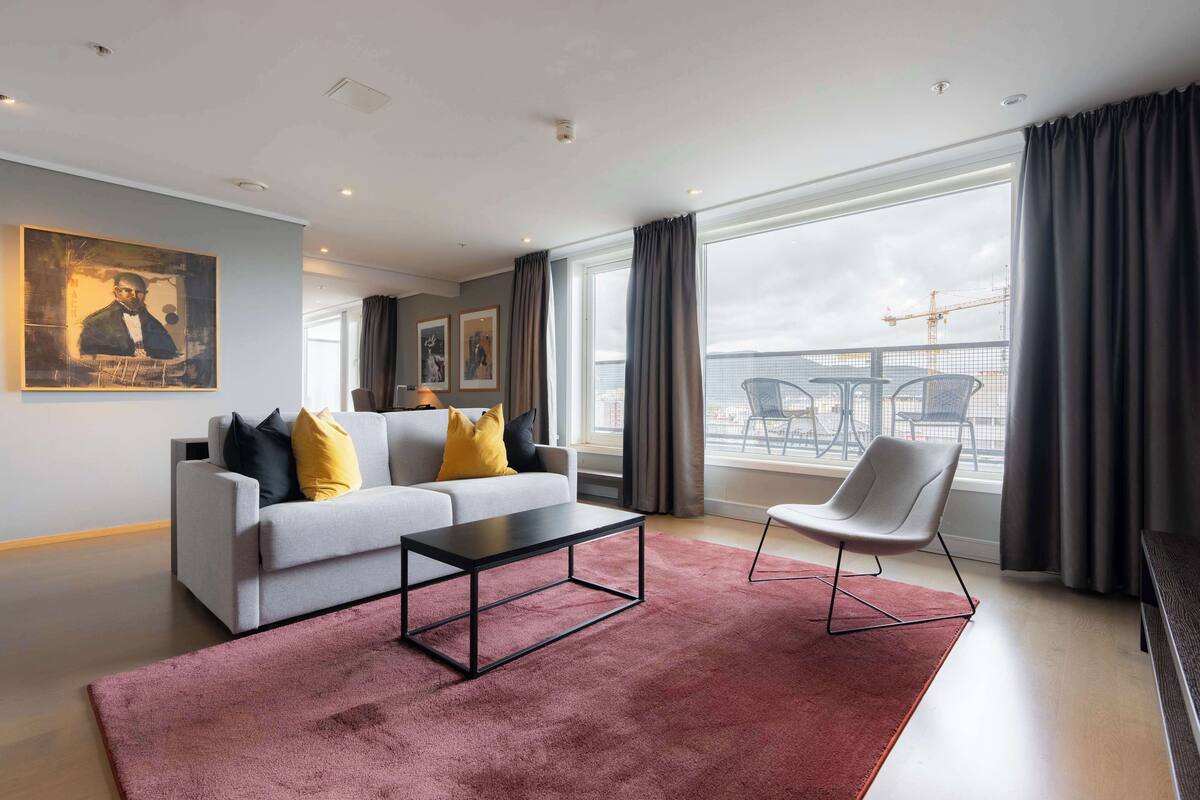 A modern living room with a grey sofa, a black coffee table, and a red rug. There are two chairs, a large window with curtains, and a balcony with a table and chairs.