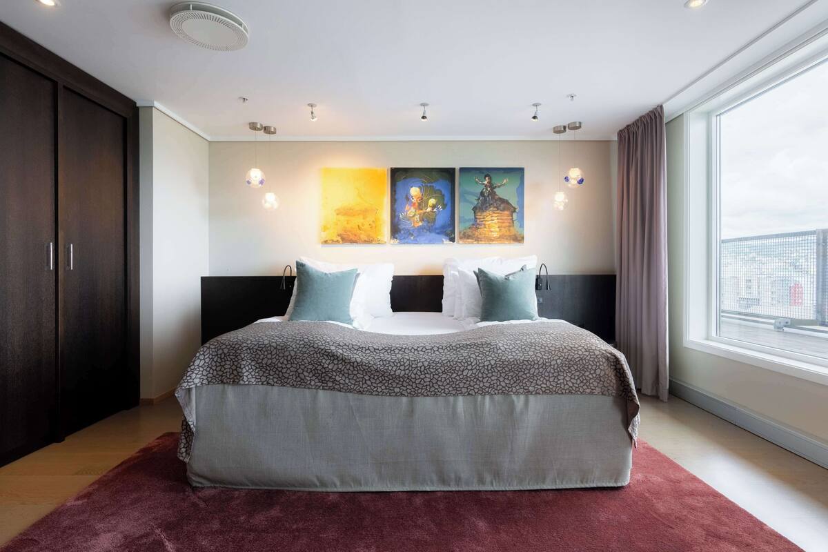 A modern bedroom with a large bed, a dark wardrobe, and a city view through a large window.