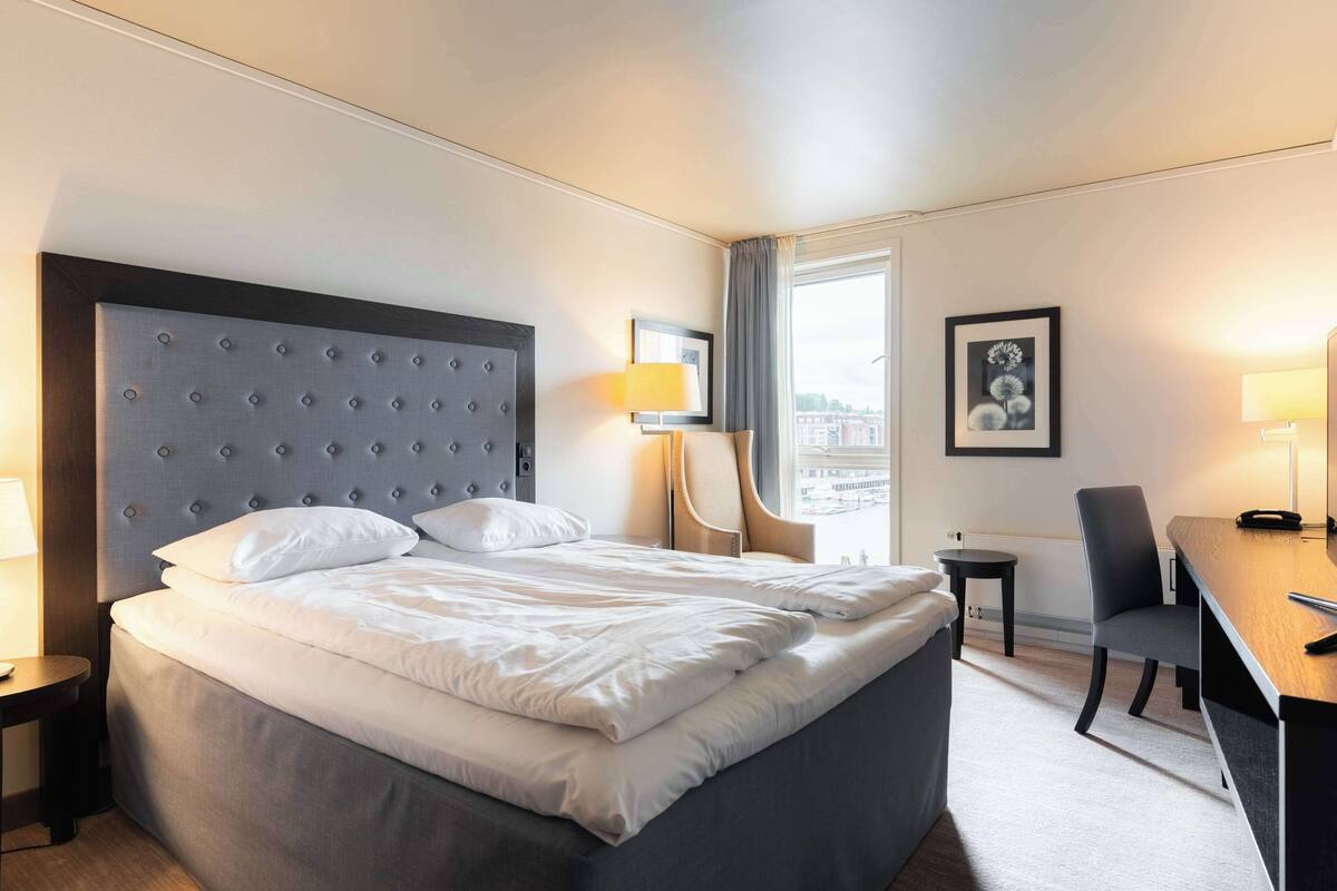 A hotel room with a large bed, a desk, a chair, a lamp, and a window with a city view.
