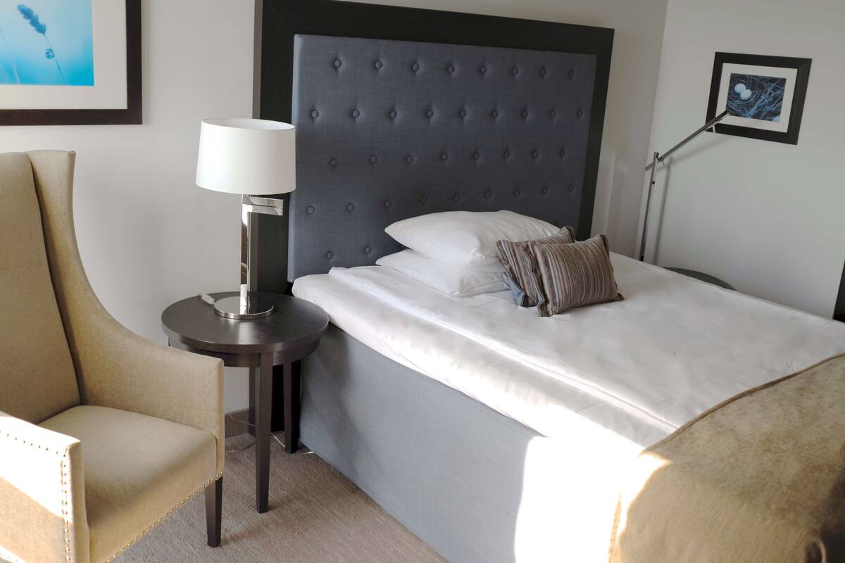 A hotel room with a tufted headboard, a round bedside table with a lamp, a chair, and a bed with pillows.