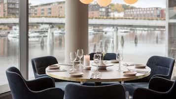 Una mesa de comedor dispuesta con platos, vasos y una vela, con vista a un río y barcos.