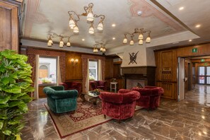Un hall d’entrée avec un sol en marbre, des fauteuils à bras en velours rouge, un fauteuil vert, une table basse ornée de fleurs et une cheminée.