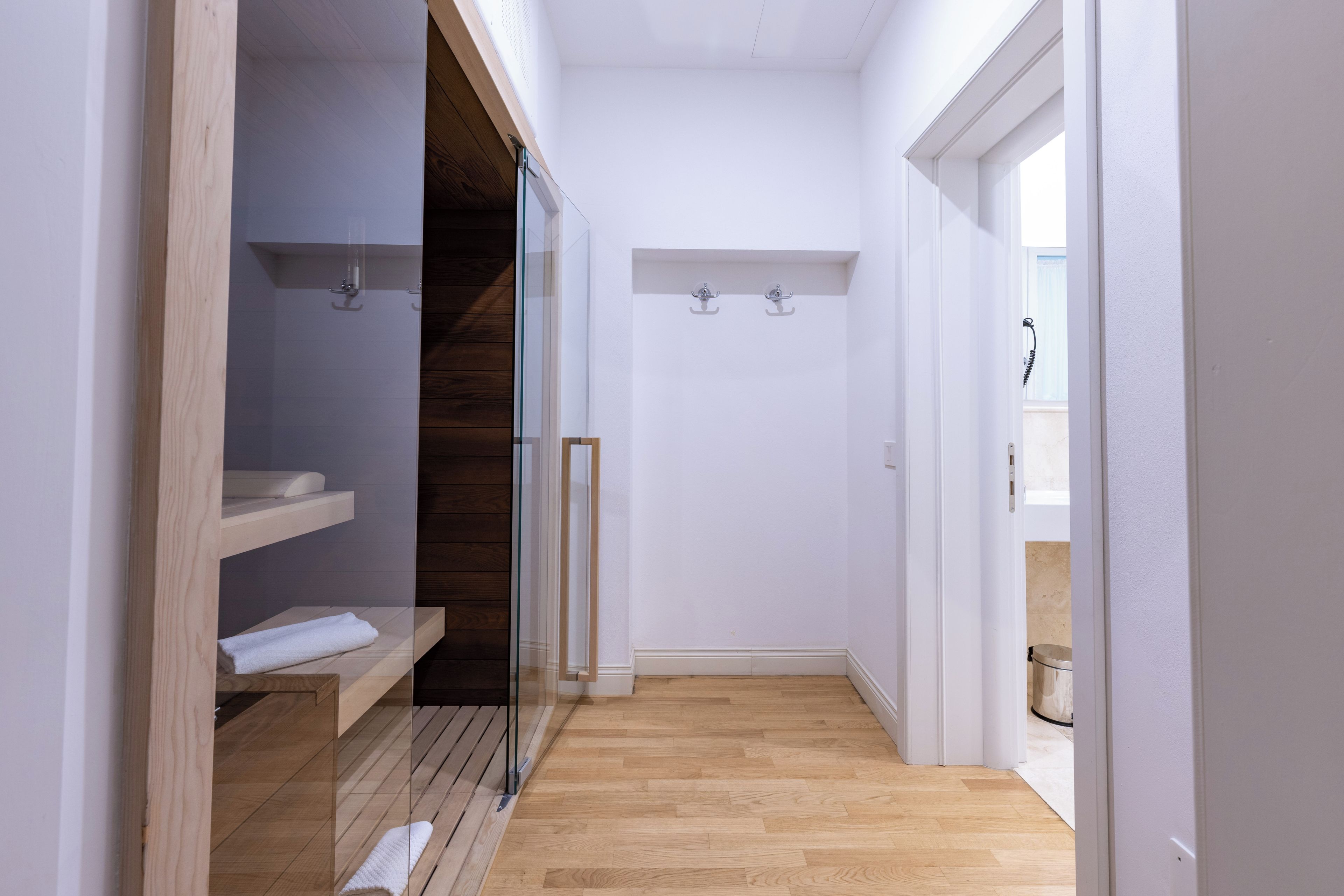 A narrow hallway with wooden flooring, a glass door leading to a room with a wooden floor, and a white wall with a small shelf.
