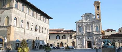 Una plaza histórica con una iglesia destacada, que cuenta con un campanario, rodeada de arquitectura clásica y un espacio abierto central con asientos y follaje.