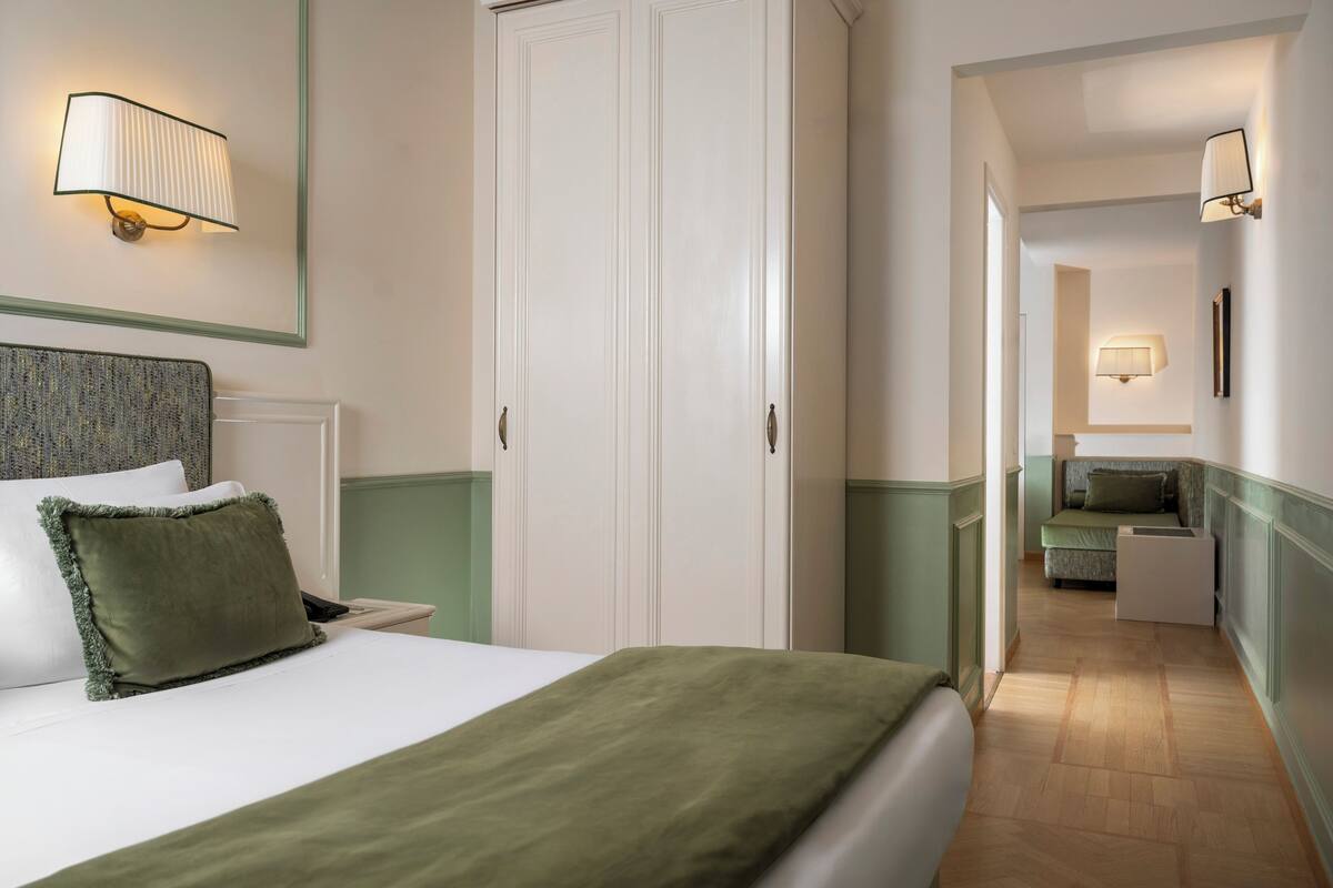 A hotel room with a bed, a wardrobe, a nightstand, a lamp, and a view into the adjacent room.