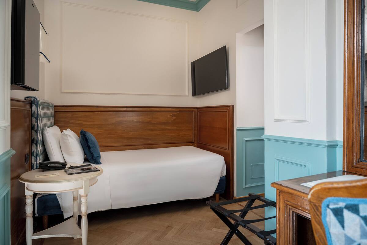 A bedroom with a wooden headboard, a single bed, a bedside table with a lamp, and a flat-screen TV mounted on the wall.